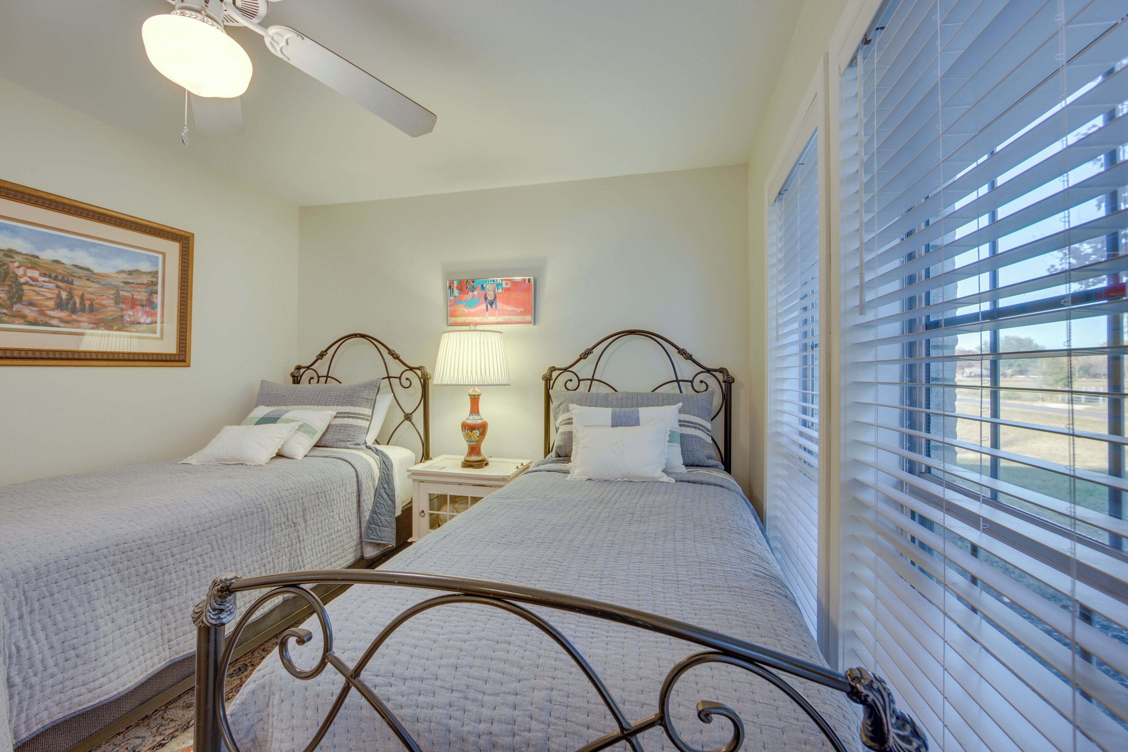 A bedroom with two beds, a ceiling fan, a nightstand with a lamp, and a window with blinds.