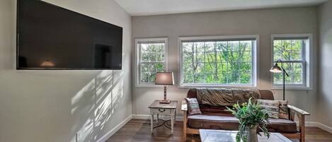 A living room with a flat-screen TV, a brown sofa, a coffee table, and a lamp. There are three windows providing natural light.