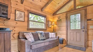 A cozy cabin interior with a wooden floor, a sofa with plaid cushions, a door with a horse logo, and a window with a view of trees. - Near Winter Trails! Rustic Cabin in Mill Spring (Mill Spring)