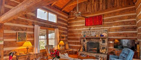 L’intérieur d’une cabane en rondins, avec une table à manger en bois, un fauteuil inclinable bleu, une cheminée en pierre et un ventilateur de plafond.