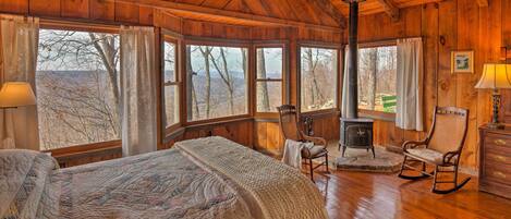 Acogedora habitación tipo cabaña con una cama grande, un sillón mecedora, chimenea y grandes ventanas que ofrecen vistas del bosque circundante.