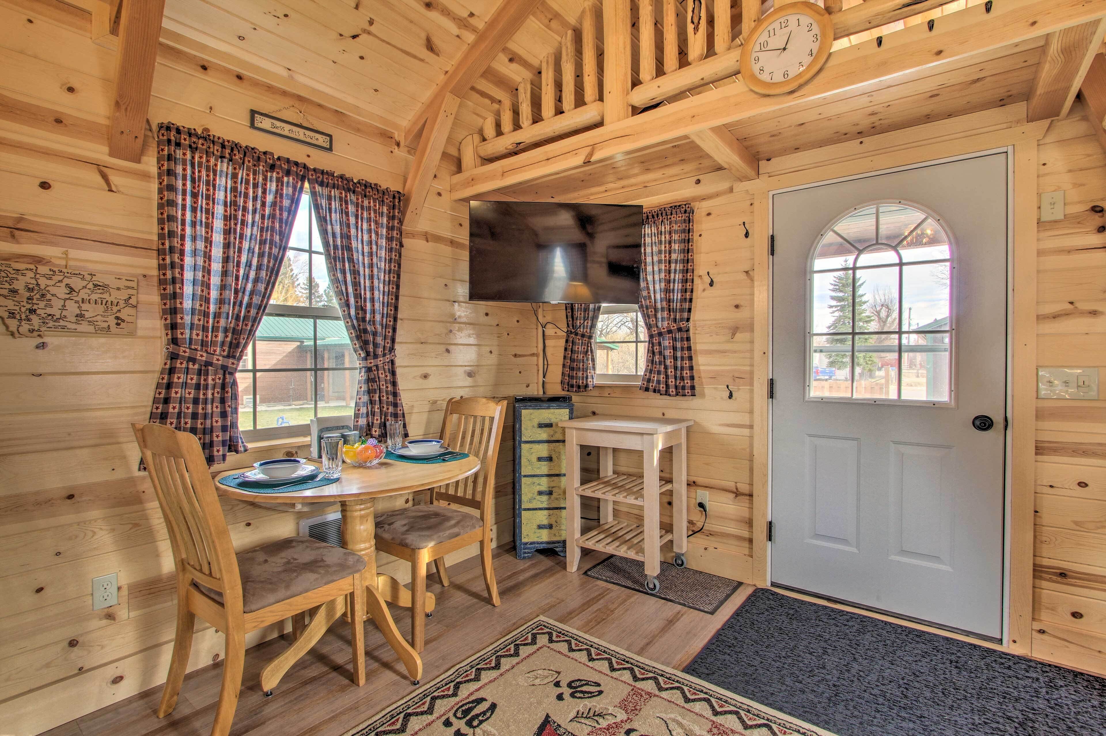 A wooden cabin interior with a dining table, chairs, a television, and a door.