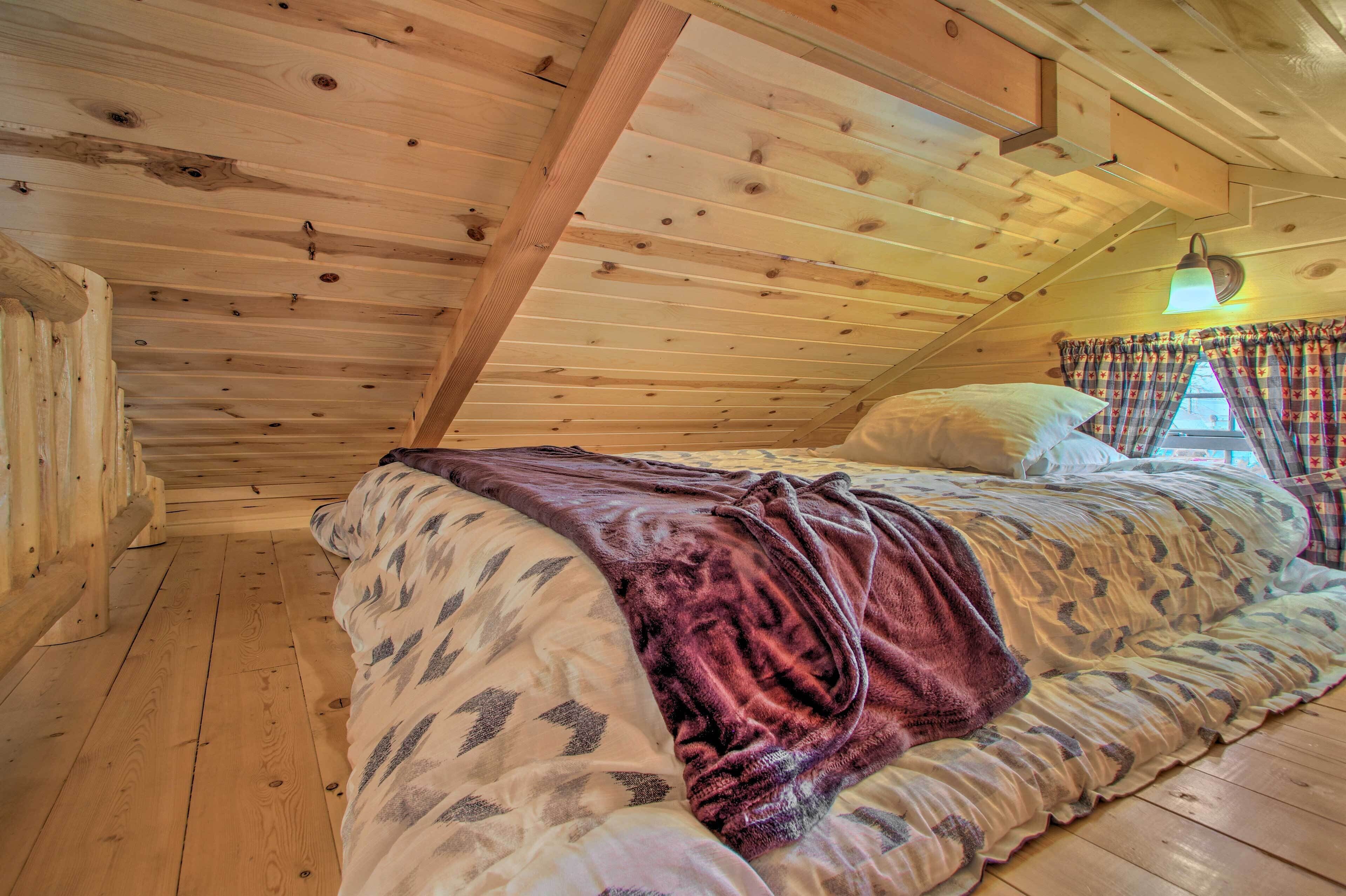 A wooden cabin bedroom with a bed, a window with curtains, and a sloped ceiling.