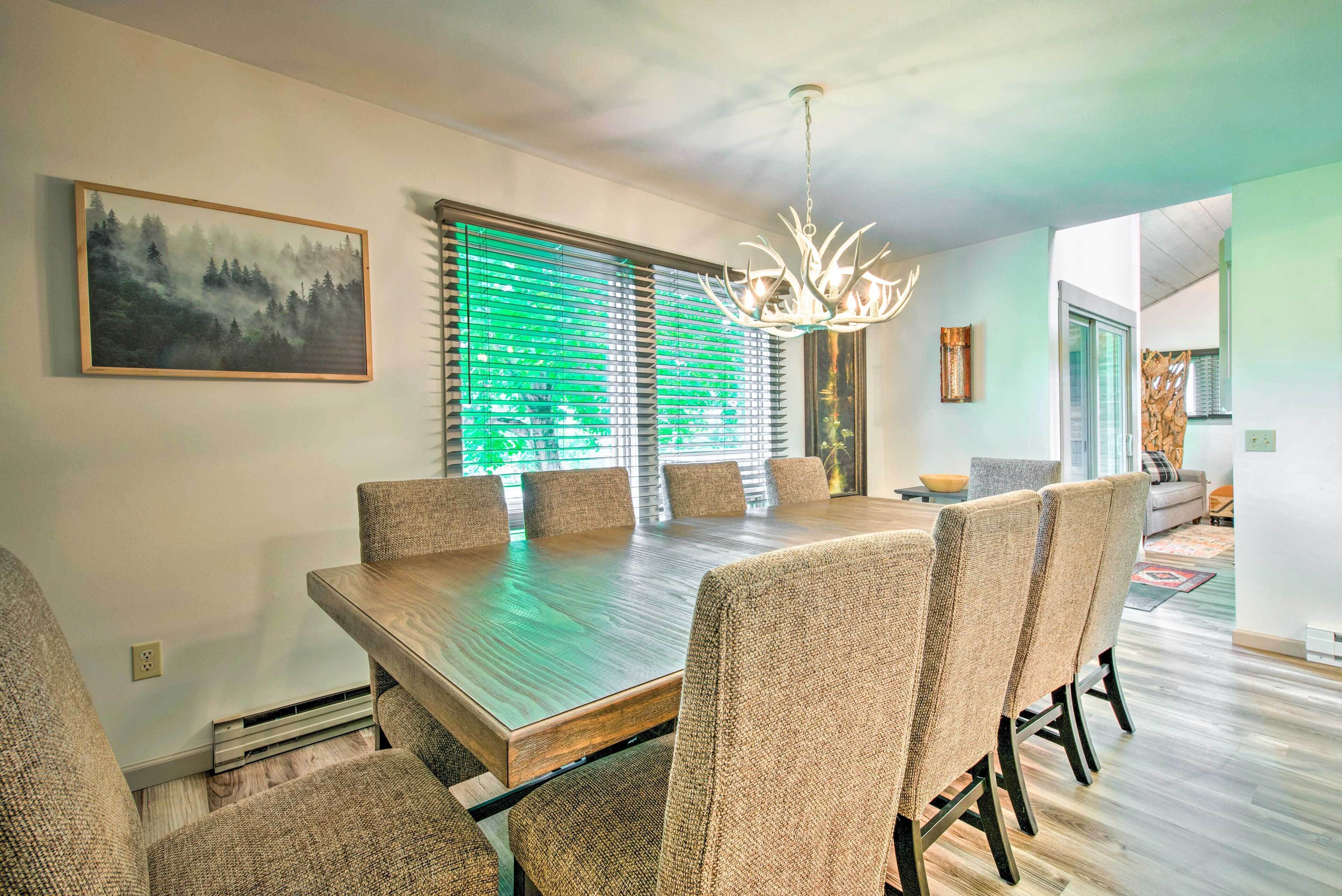 A dining area with a large wooden table and six upholstered chairs, a chandelier, and a painting on the wall.