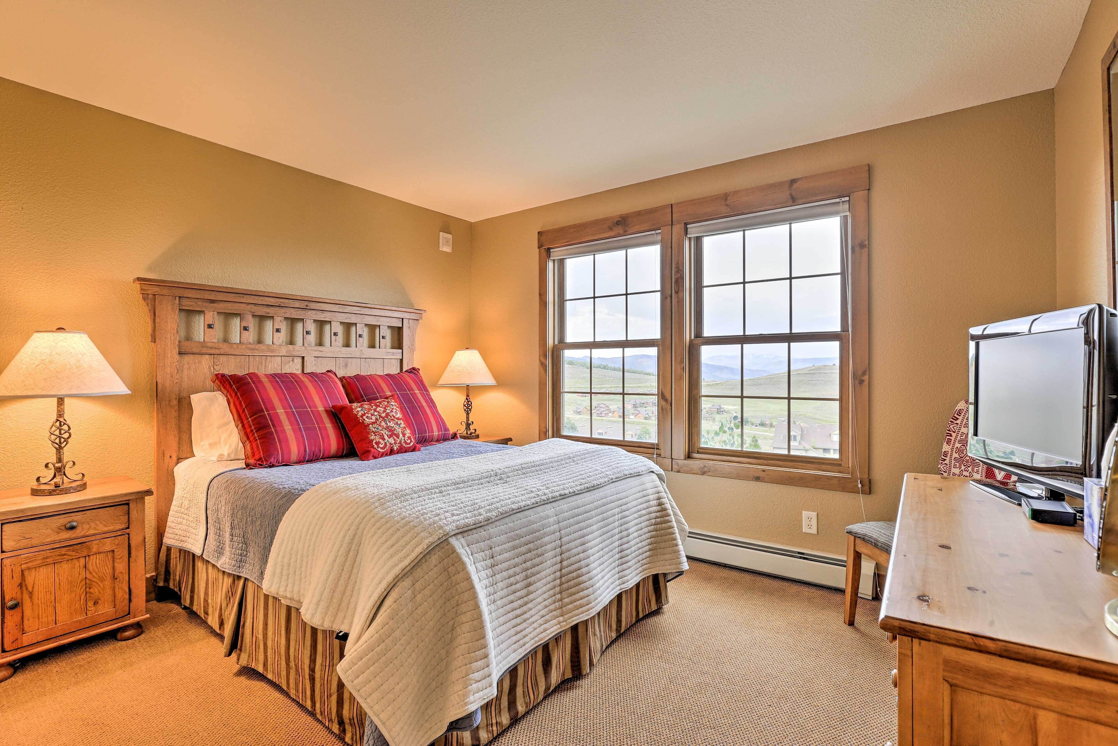 A bedroom with a large bed, two bedside lamps, a wooden headboard, a dresser, a television, and a window with a view of the outdoors.