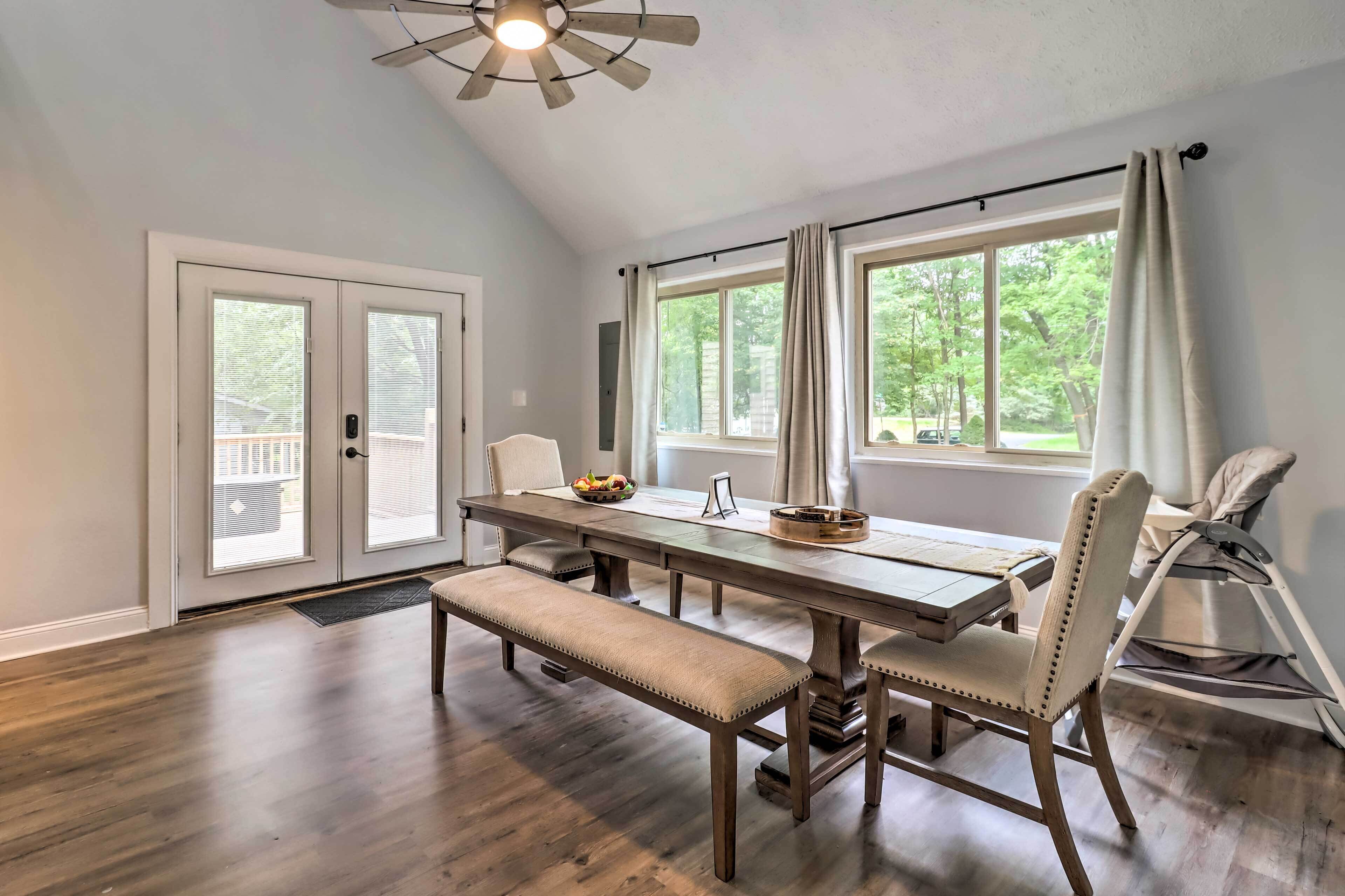 A dining area with a wooden table, bench, and chairs, large windows with curtains, and a ceiling fan.