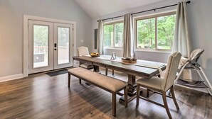 Un espace repas comprenant une table, un banc et des chaises en bois, de grandes fenêtres avec des rideaux et un ventilateur de plafond.
