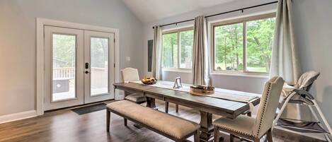 A dining area with a wooden table, bench, and chairs, large windows with curtains, and a ceiling fan.