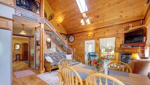 A cozy cabin interior with a dining area, wooden furniture, a fireplace, and a staircase leading to an upper level.