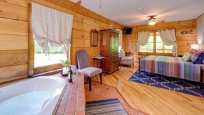 A wooden cabin interior with a bed, a chair, a table with a bottle and glasses, a cabinet, and a ceiling fan.