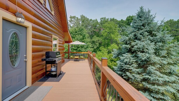 A wooden deck with a grill and outdoor furniture, surrounded by lush greenery and a large evergreen tree.