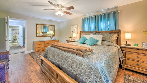 A bedroom with a large bed, a ceiling fan, a nightstand with a lamp, and a wooden dresser.