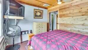 Een slaapkamer met een bed, een ventilator, een televisie op een standaard, een houten kast en een plafondventilator.