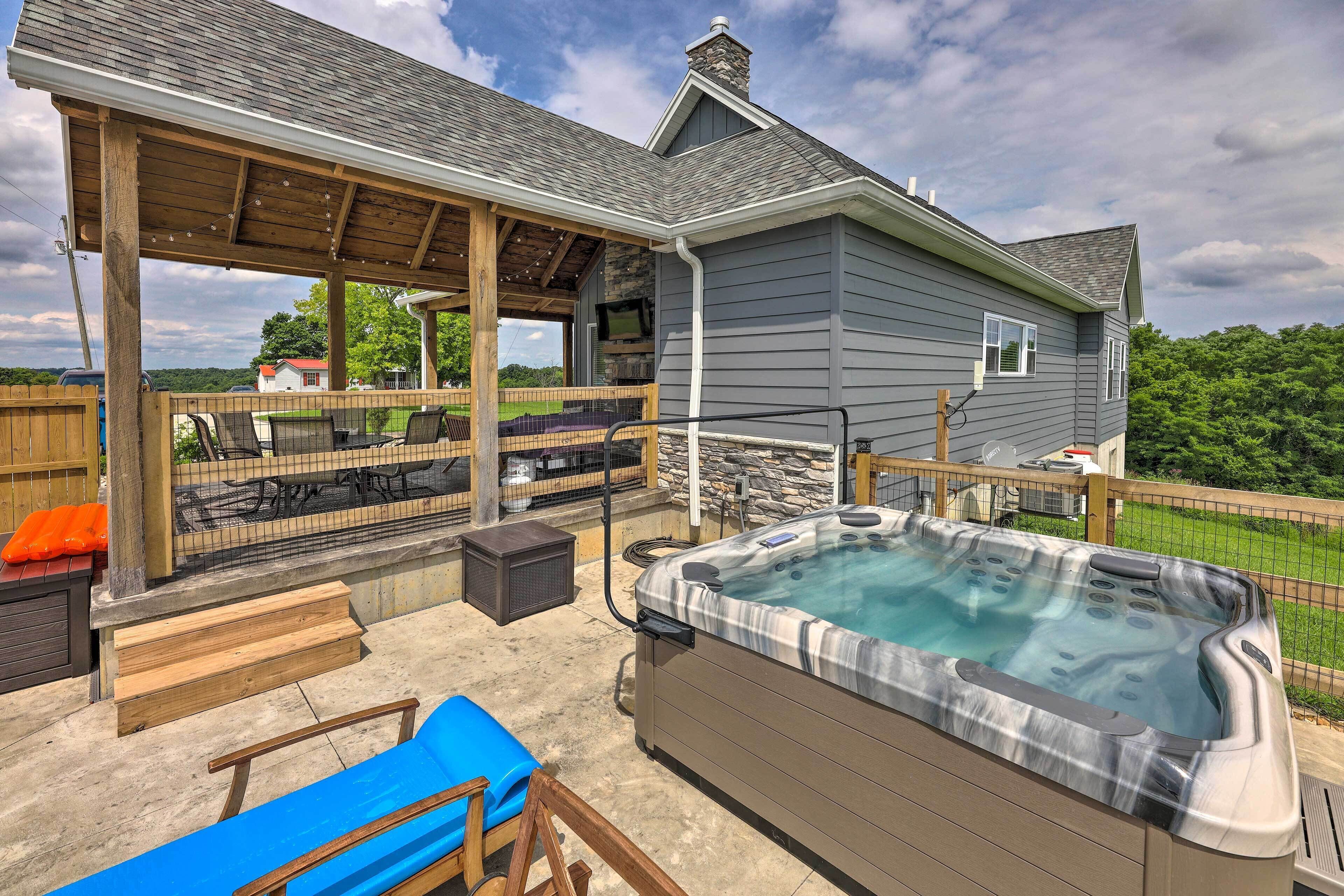 A covered outdoor patio with a hot tub, seating, and a stone fireplace.