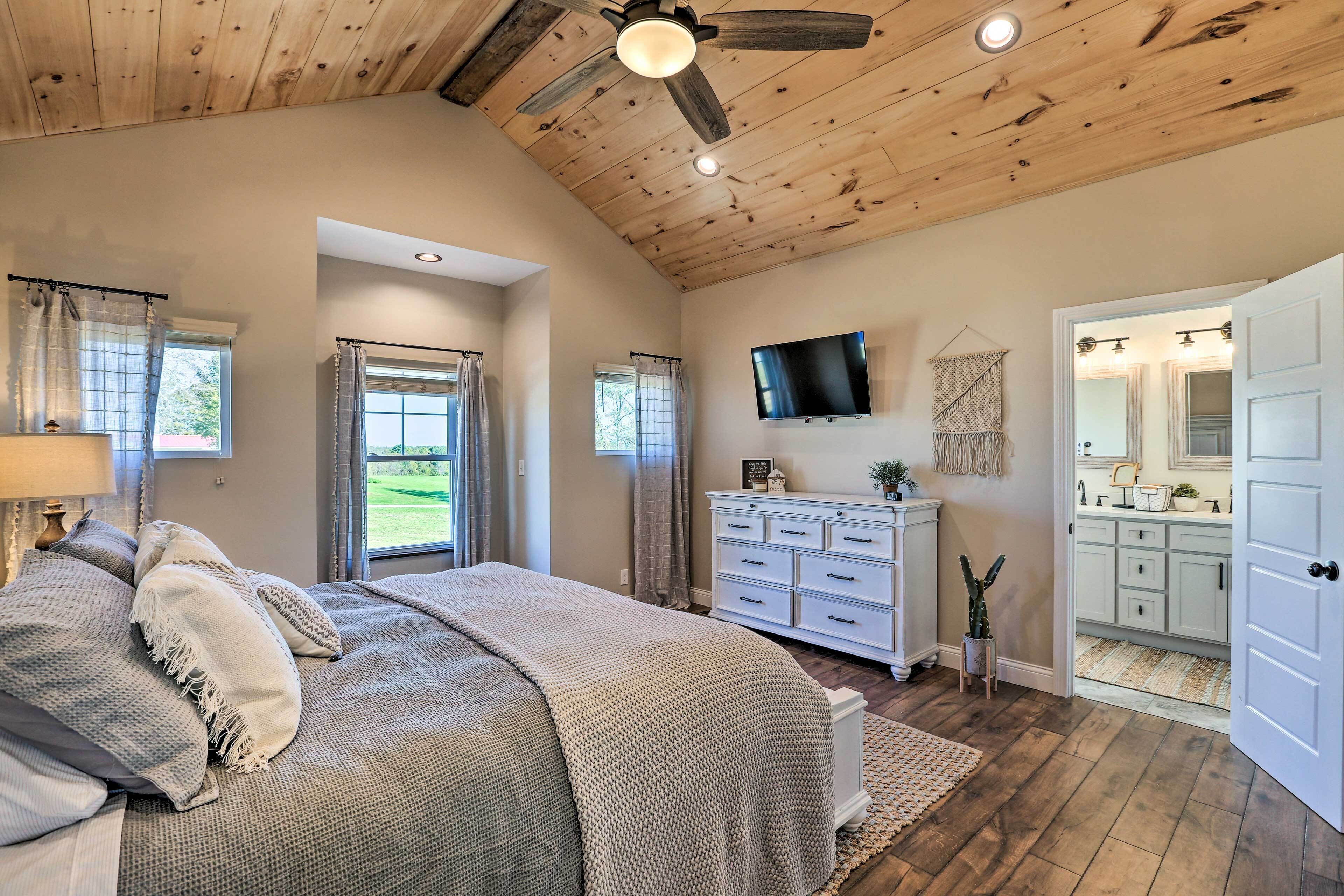 A bedroom with a large bed, a ceiling fan, a TV, a dresser, and a view of the bathroom.