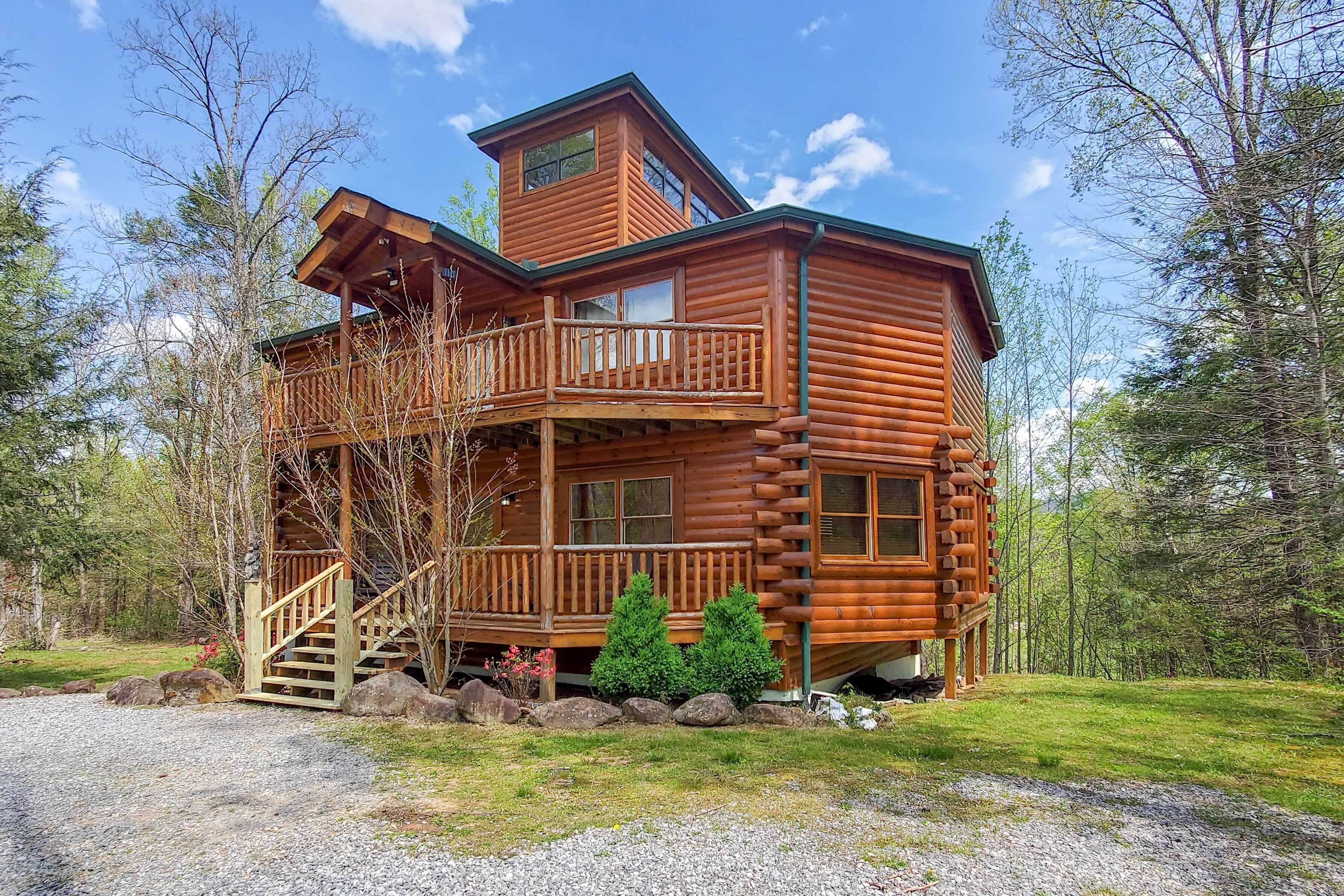 A log cabin with a balcony and a second-story window, surrounded by trees and a gravel driveway.