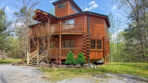 Une cabane en rondins dotée d’un balcon et d’une fenêtre au deuxième étage, entourée d’arbres et bordée d’une allée en gravier.
