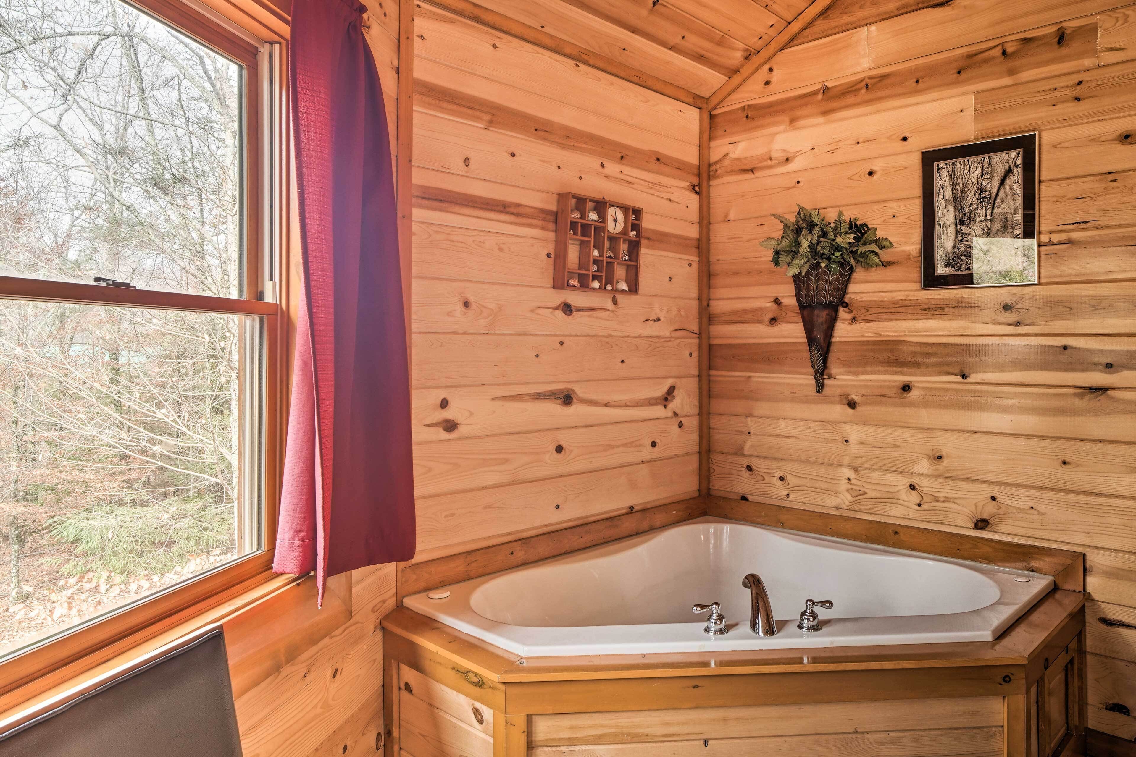A wooden cabin bathroom with a large bathtub, a window with a view of trees, a clock, and a framed picture on the wall.