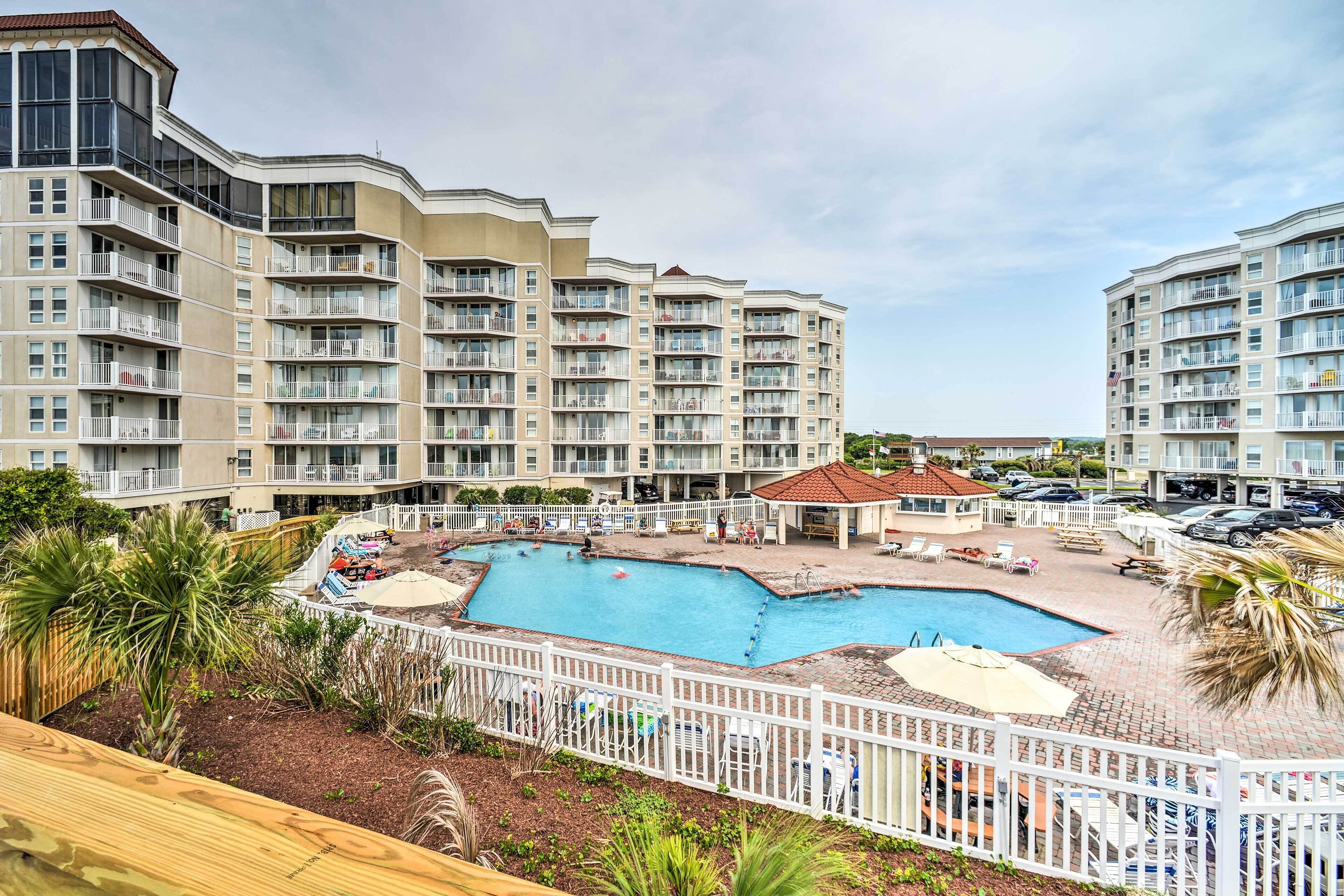 A multi-story hotel with a swimming pool surrounded by a white fence, palm trees, and lounge chairs.