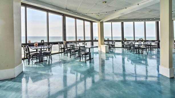 A spacious indoor area with large windows overlooking the sea, featuring multiple tables and chairs arranged for dining or meetings. - Colorful Beachfront Condo w/ Resort Amenities (Sneads Ferry)