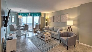 A living room with a sofa, armchair, coffee table, and dining area. There is a TV mounted on the wall, a lamp, and artwork on the wall. - Colorful Beachfront Condo w/ Resort Amenities (Sneads Ferry)