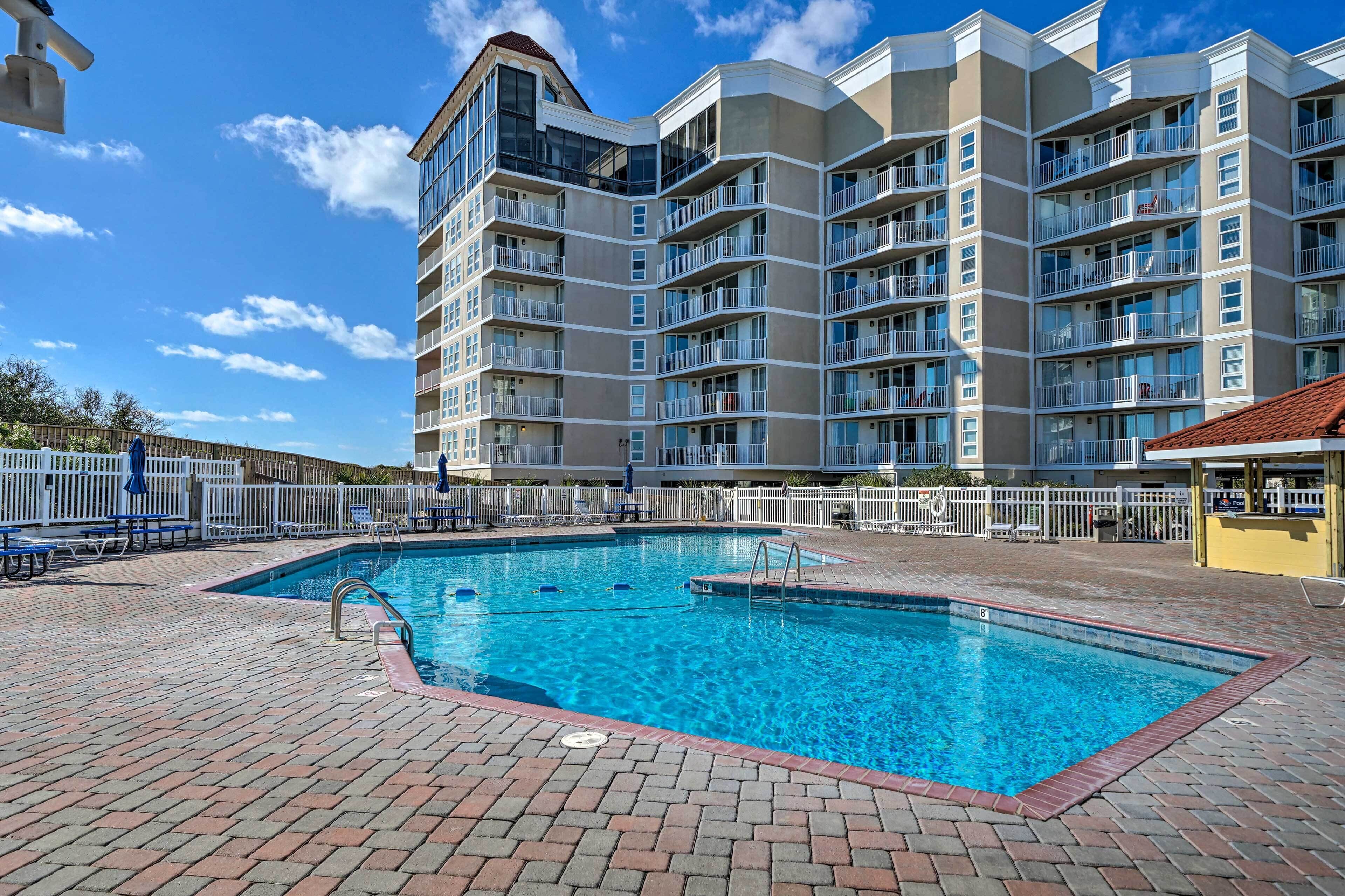 A swimming pool with a brick deck, surrounded by a white fence, and a multi-story building with balconies in the background.