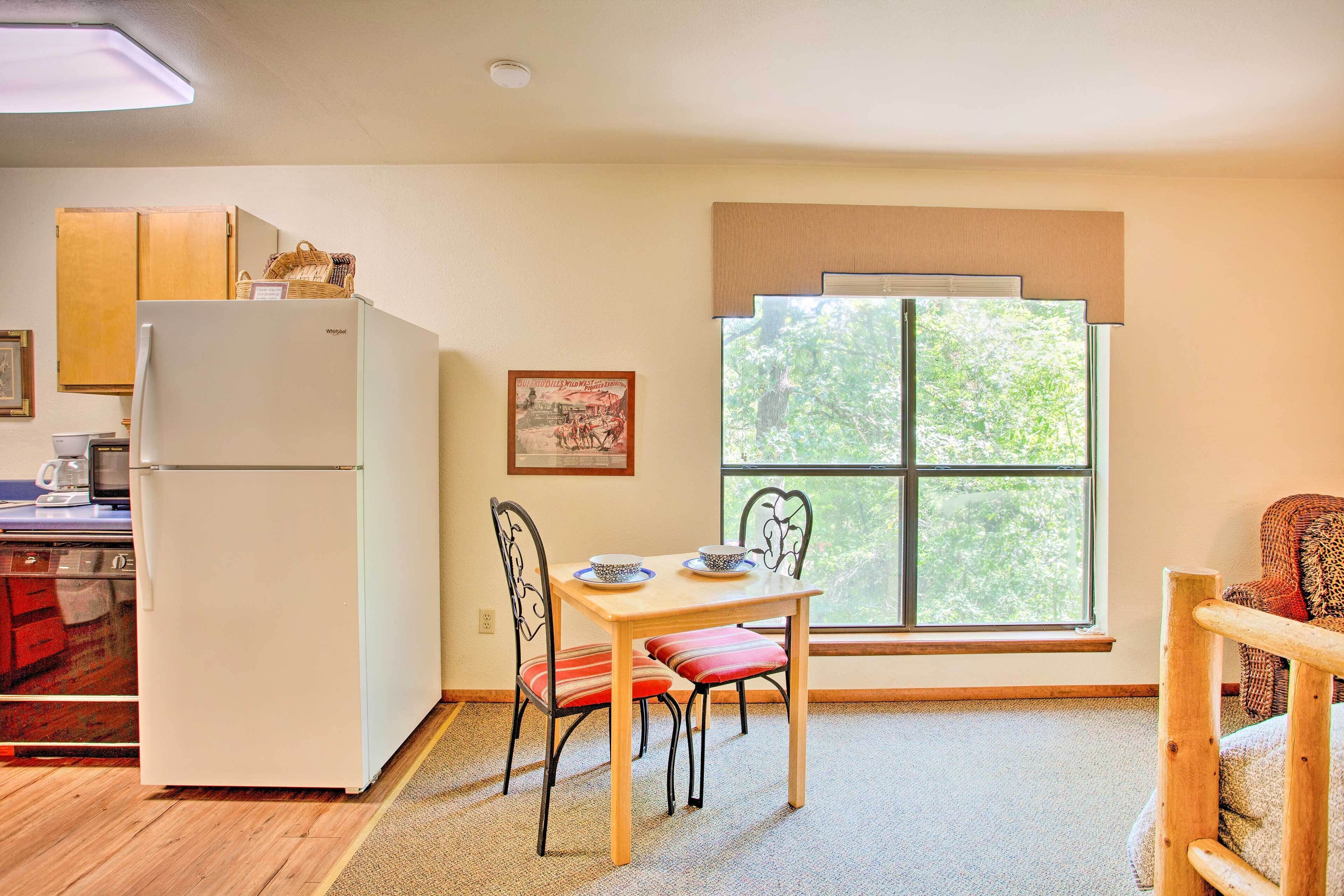 Cocina compacta con refrigerador, una mesa de comedor con sillas, una ventana con persianas y vista a la vegetación exterior.