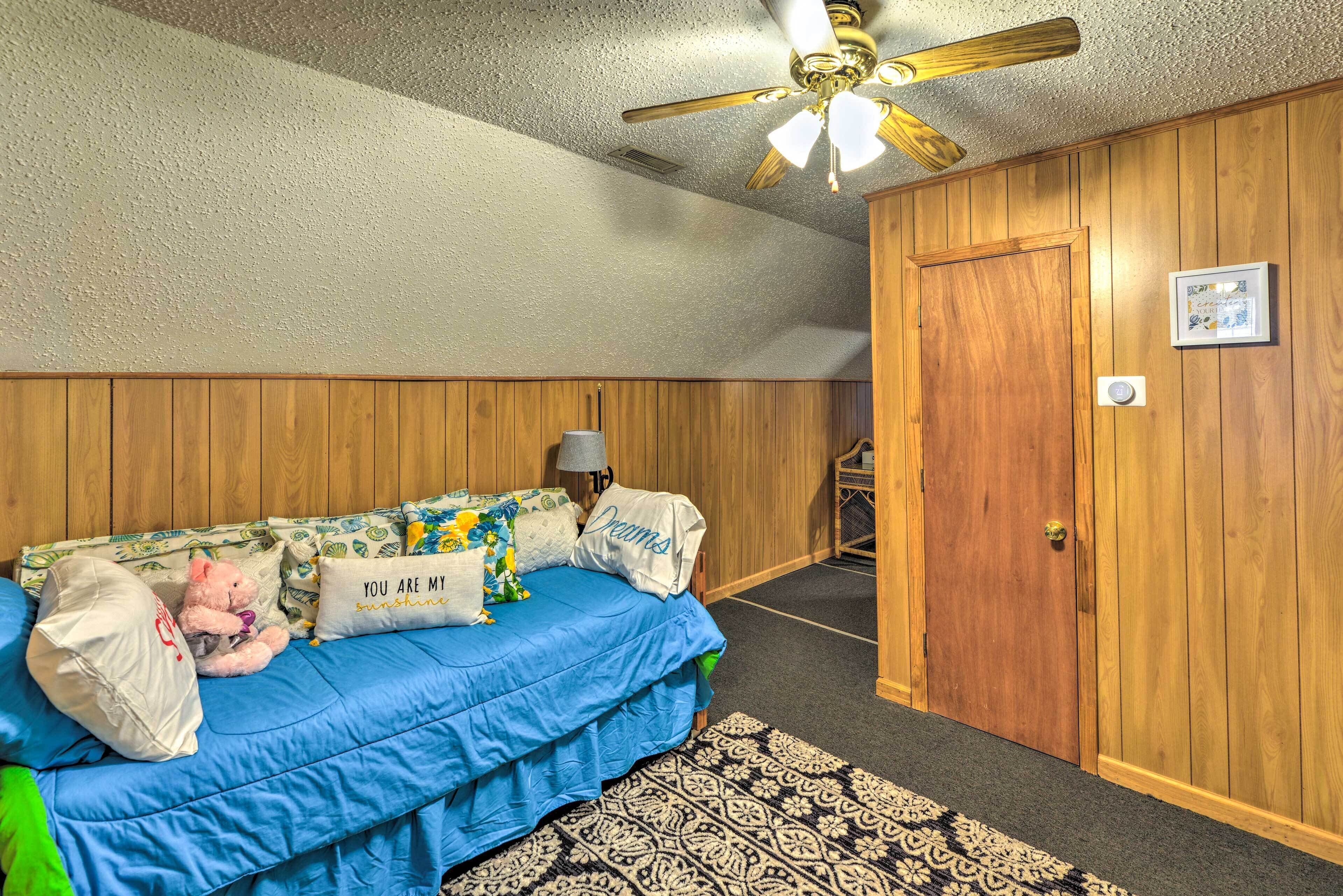 A bedroom with a blue bed, wooden walls, a ceiling fan, and a patterned rug.
