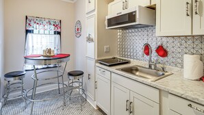 Una cucina con mobili bianchi, un lavello in acciaio inossidabile, un forno a microonde e una parete rivestita di piastrelle. Sono presenti due sgabelli e un piccolo tavolo con piano rosso.