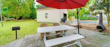 A picnic table with a red umbrella, a brick patio, and a small house in the background.