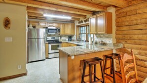 A kitchen with wooden cabinets, a stainless steel refrigerator, a granite countertop, and a wooden chair.