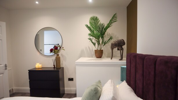 A bedroom with a bed, a dresser, a mirror, a plant, and a statue.