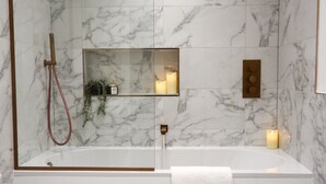 A modern bathroom with a marble wall, a glass-enclosed bathtub, a handheld showerhead, a shelf with candles, and a towel rack.