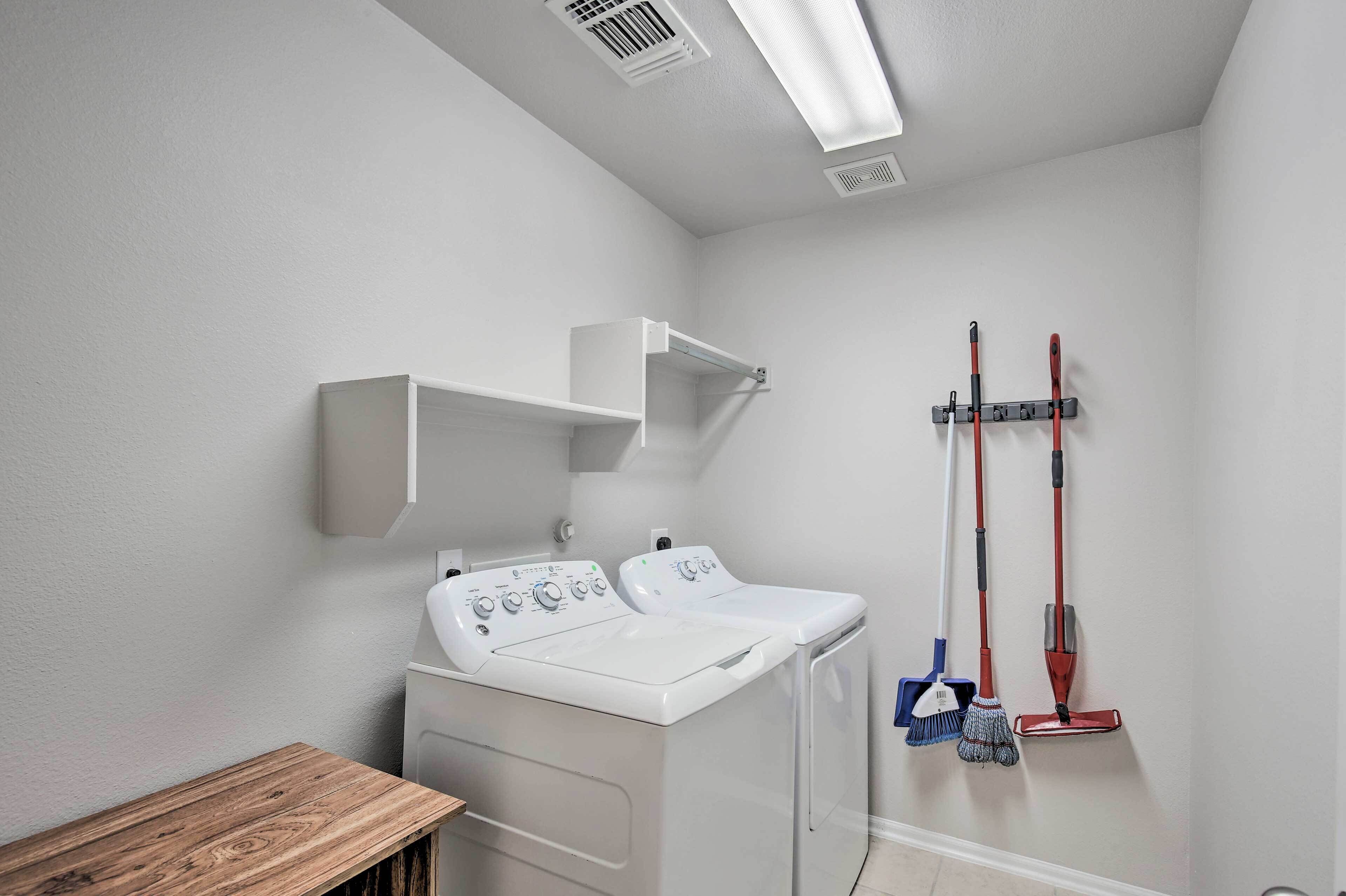 Une buanderie équipée de deux machines à laver à chargement frontal blanches, d’un banc en bois et d’un étendoir mural agrémenté de deux balais.