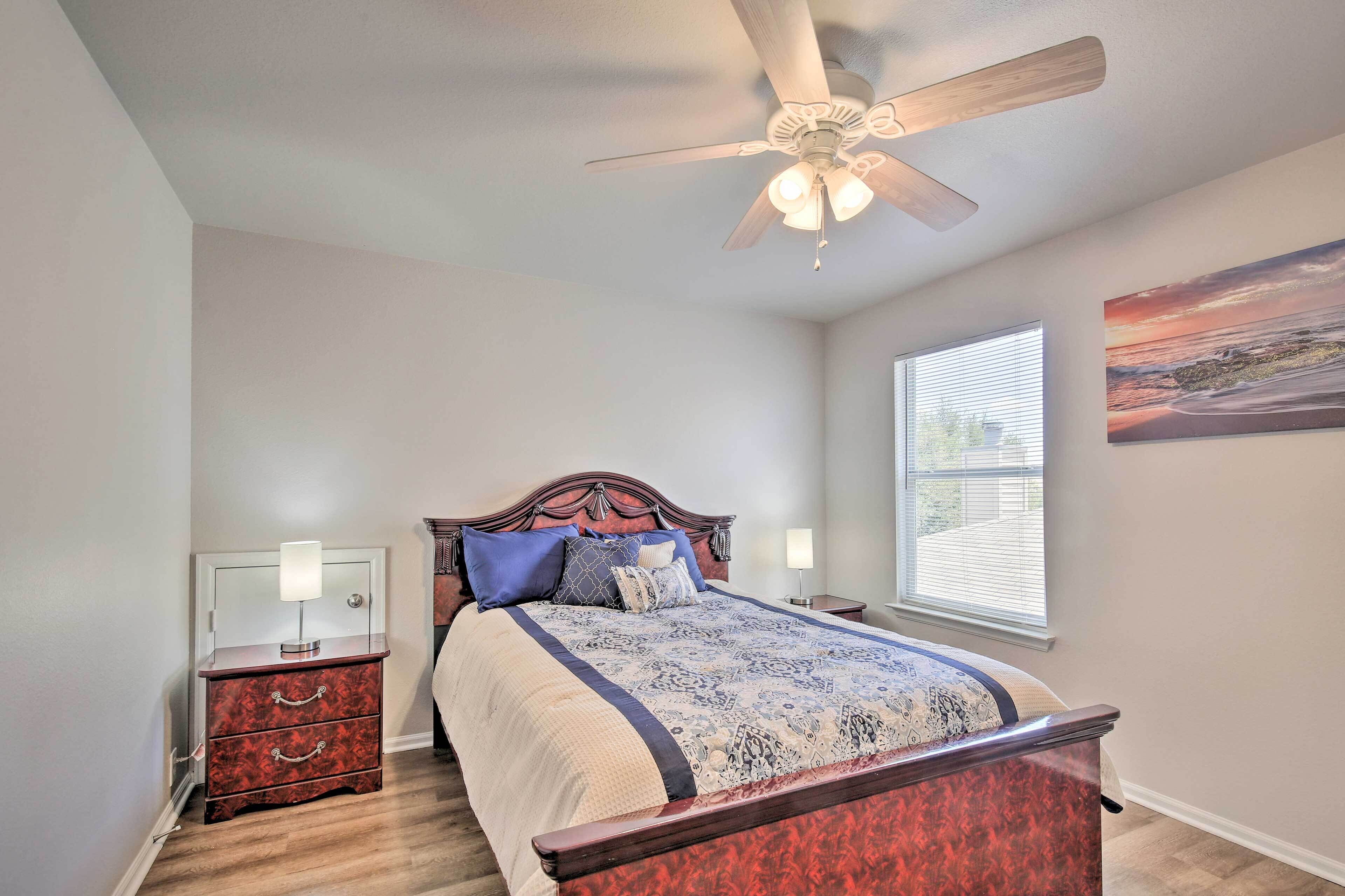 A bedroom with a bed, nightstands, a ceiling fan, a window with blinds, and a painting on the wall.