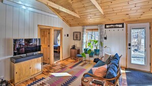 A cozy cabin interior with a wooden ceiling, a television on a cabinet, a sofa with cushions, and a door leading outside. - Hiking Trails On-site: Cozy Falls Brook Cabin (Brattleboro)