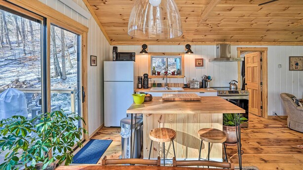 A kitchen with wooden cabinets, a central island, and a large window overlooking a snowy landscape. - Hiking Trails On-site: Cozy Falls Brook Cabin (Brattleboro)