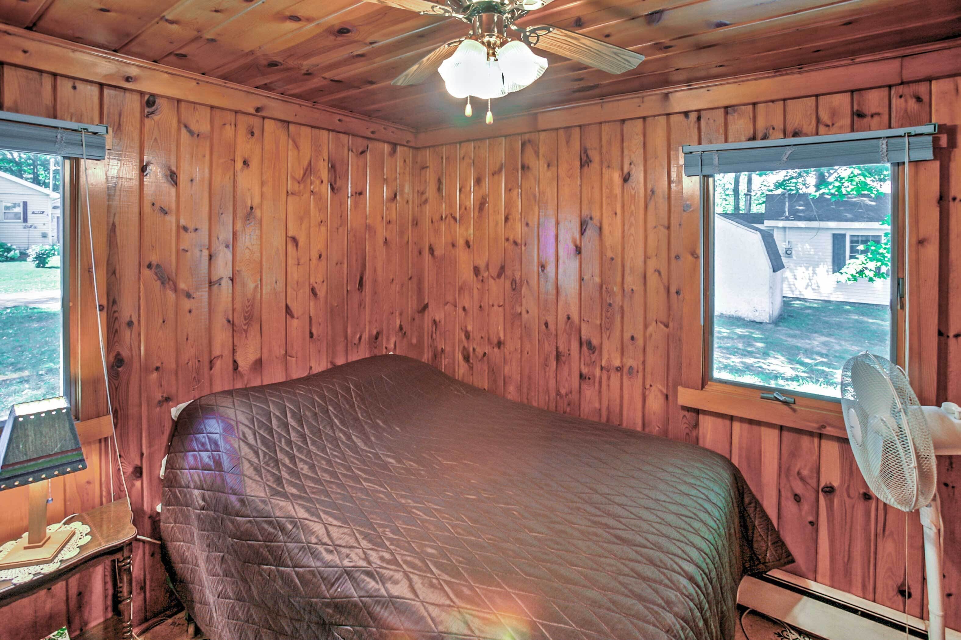 Une chambre avec des murs en bois, un lit, un ventilateur de plafond et une fenêtre donnant sur une maison et de la verdure.