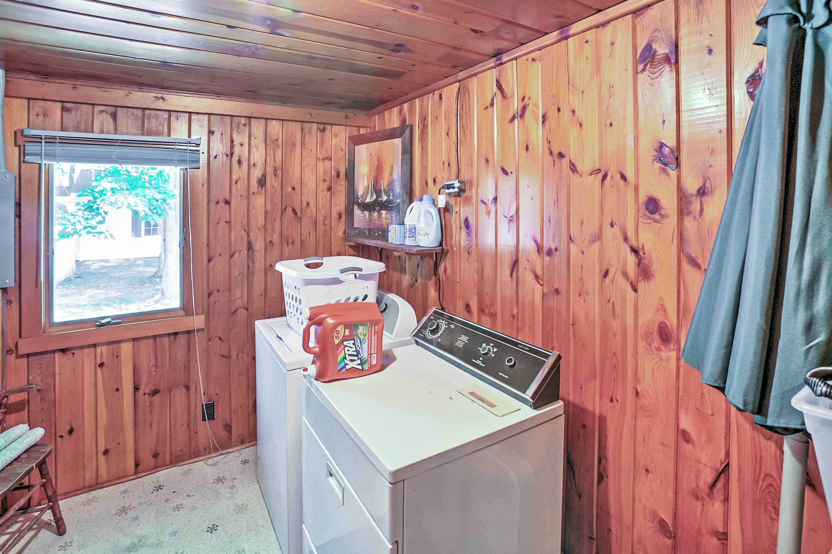 A compact laundry room with wooden walls, a washing machine, a dryer, a window with a view of greenery, and a shelf with a painting and bottles.