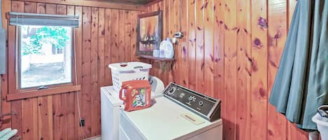 A compact laundry room with wooden walls, a washing machine, a dryer, a window with a view of greenery, and a shelf with a painting and bottles.