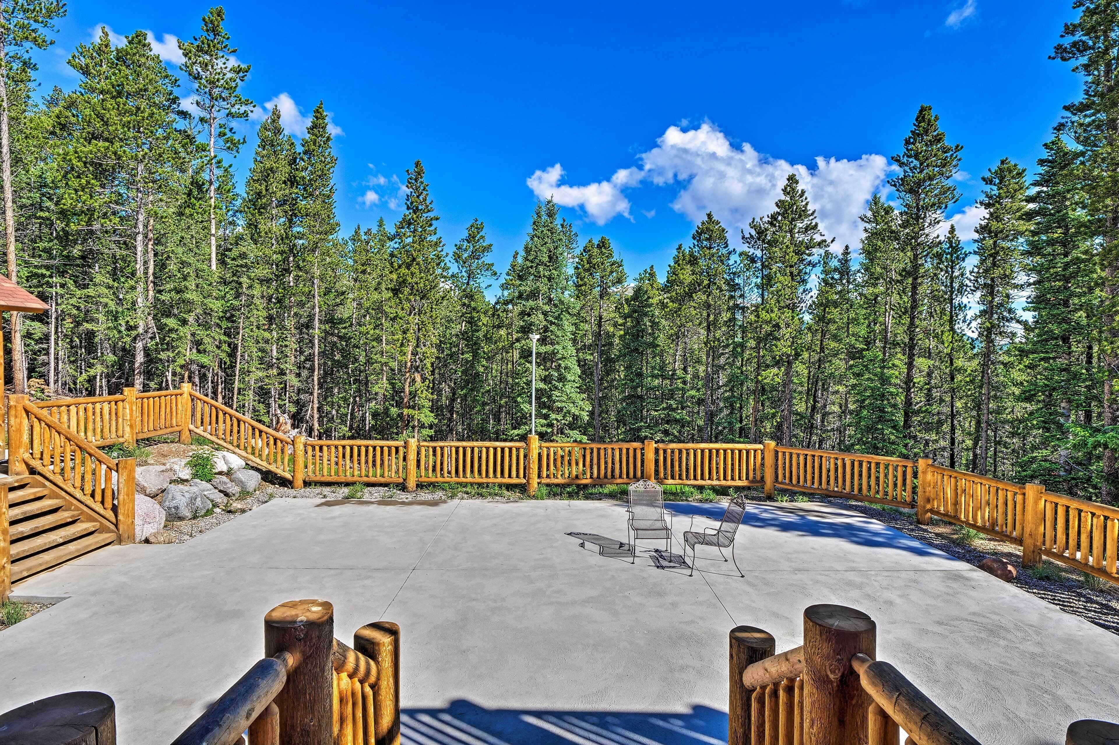Une terrasse en béton avec des garde-corps en bois, entourée d’une forêt de grands pins sous un ciel bleu dégagé.