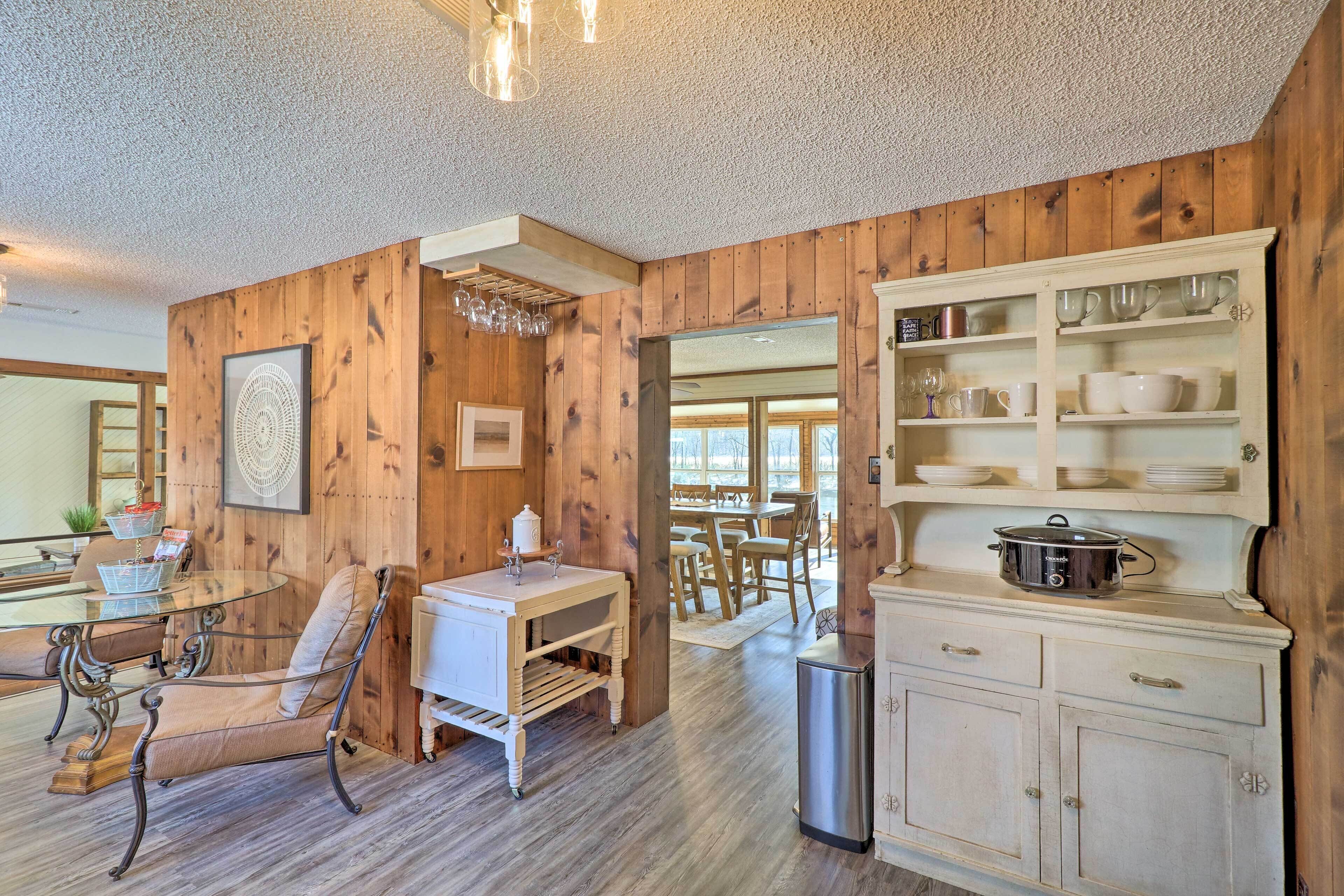 A wooden-paneled room with a dining table, chairs, a glass-top table, a white cabinet with dishes, and a trash can.