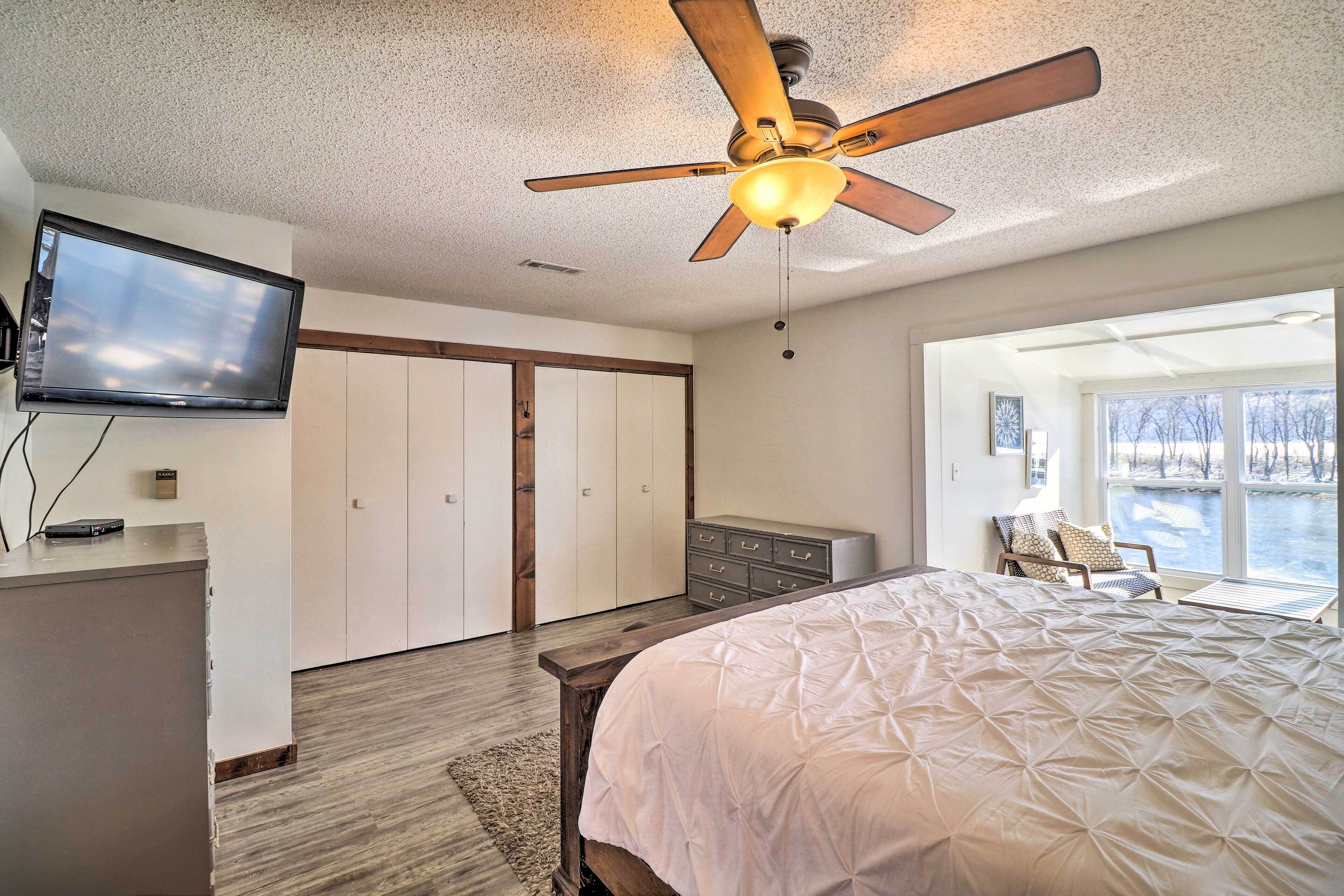A bedroom with a bed, a ceiling fan, a TV, a dresser, a chair, and a view of a lake through a large window.