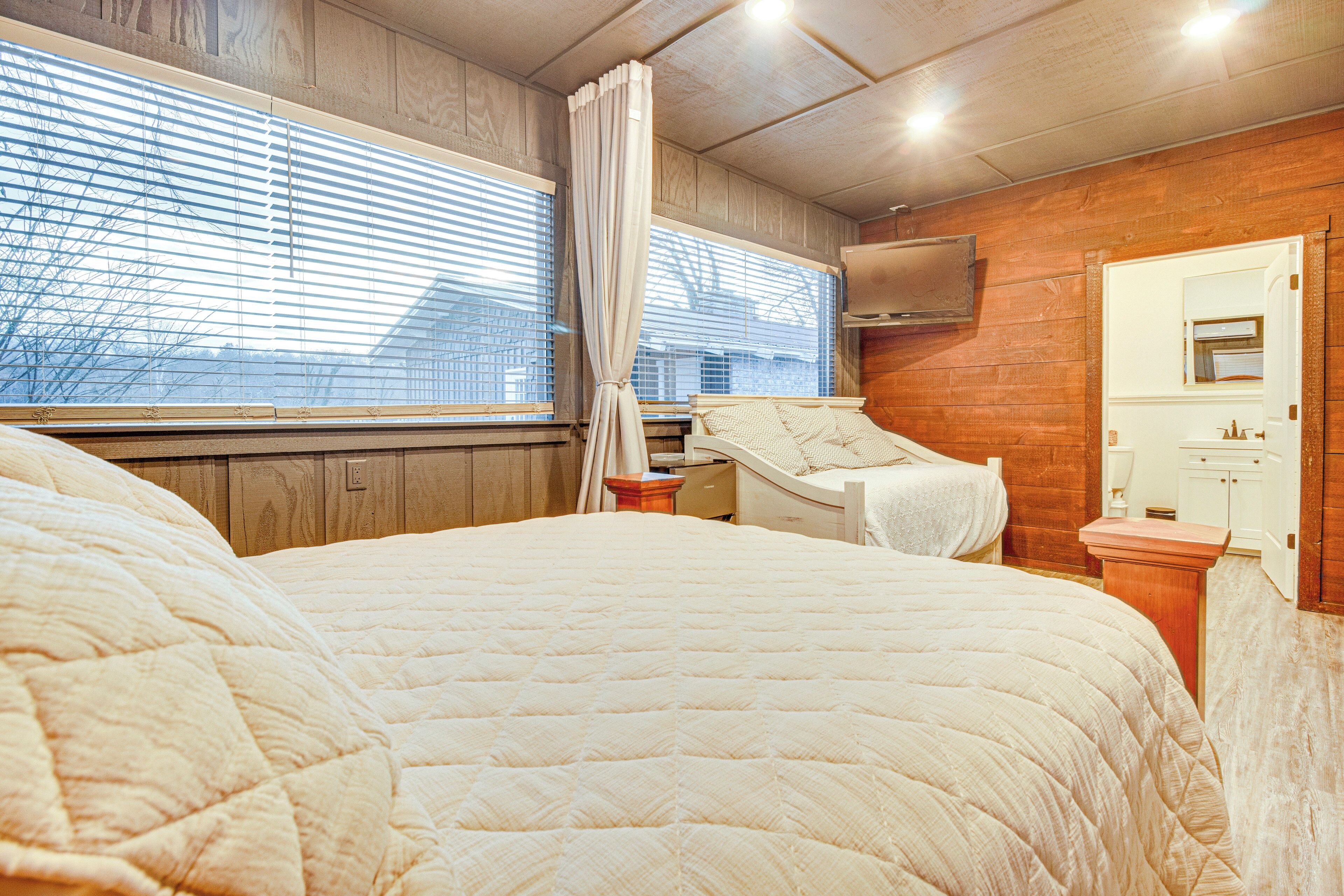 A bedroom with a large bed, a window with blinds, a wooden wall, and a mirror.