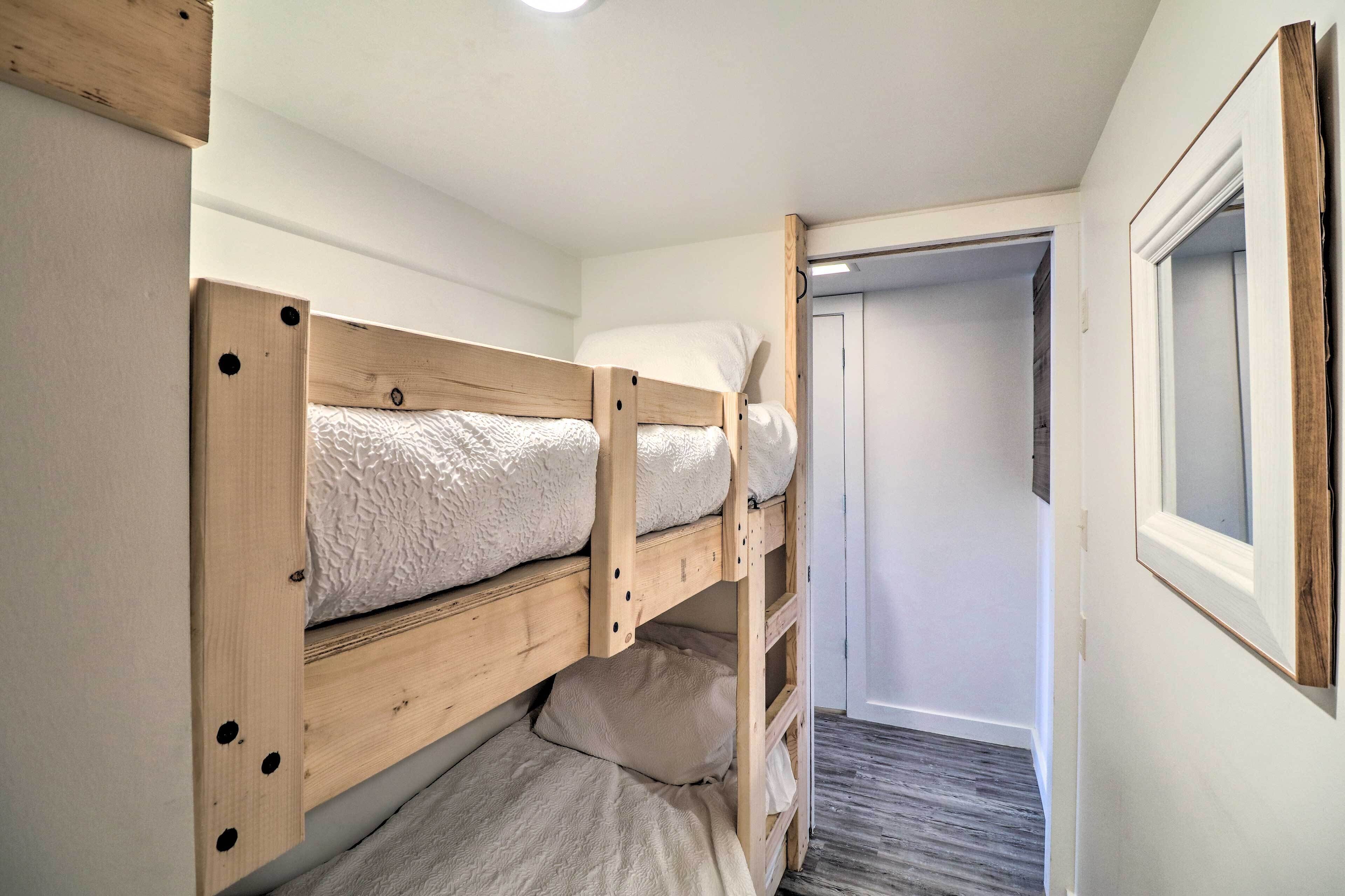Una cama litera con estructura de madera y colchón blanco. Se ve una puerta que da a otra habitación.