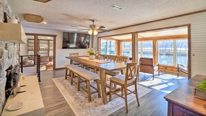 A spacious dining area with a wooden table and benches, large windows with a view of a body of water, and a stone fireplace. - 'fish House' w/ Deck, Fire Pit & River Views (Cotter)
