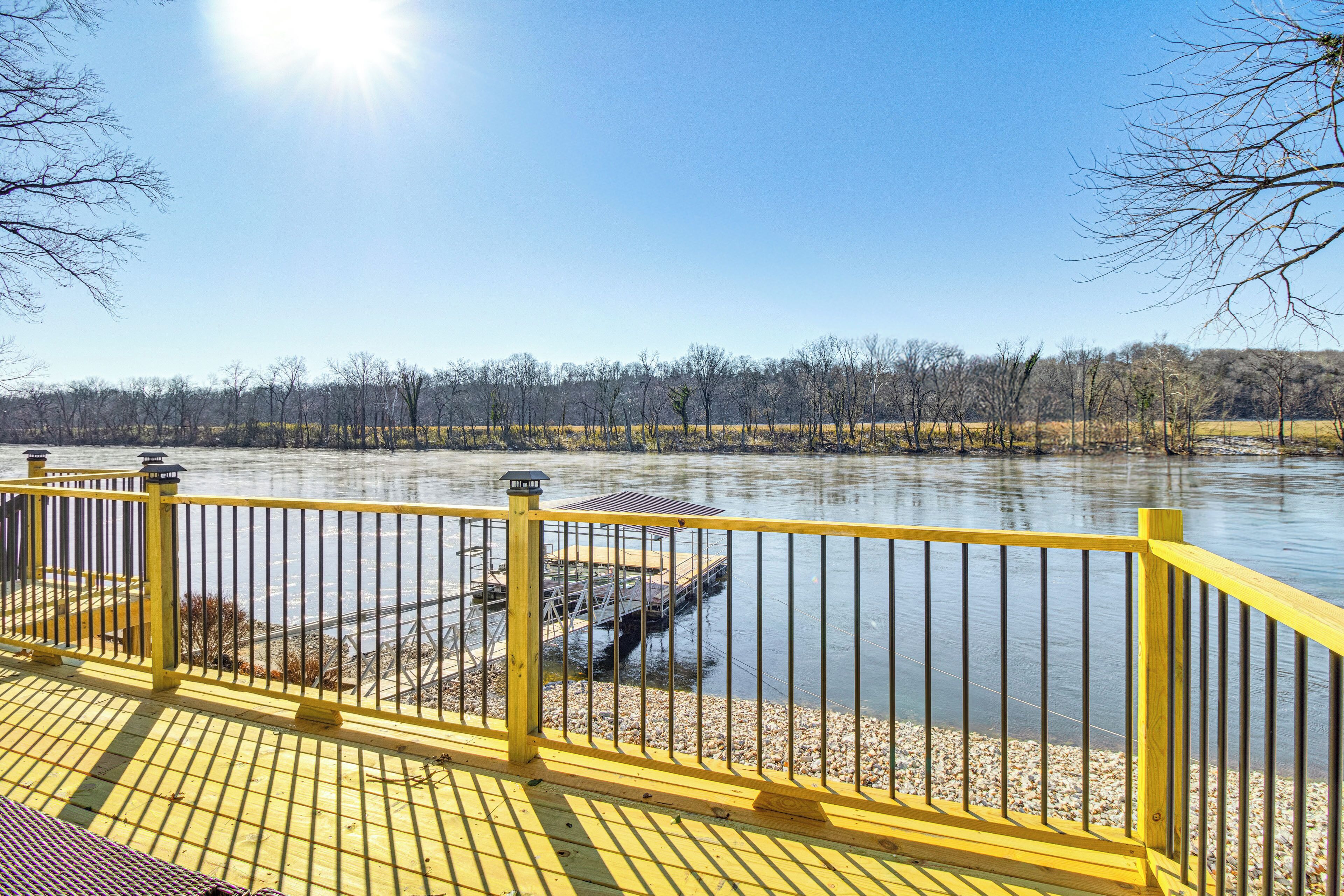 A wooden deck with yellow railings overlooking a calm body of water, a dock, and a forested area in the background.