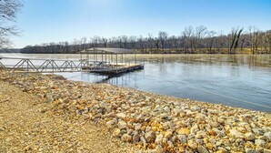 A rocky shoreline with a dock extending into a calm body of water, surrounded by leafless trees and a clear sky. - 'fish House' w/ Deck, Fire Pit & River Views (Cotter)