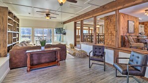 A spacious living area with a large sofa, a coffee table, and two chairs. There are ceiling fans, a fireplace, and a view of the outside through large windows. - 'fish House' w/ Deck, Fire Pit & River Views (Cotter)