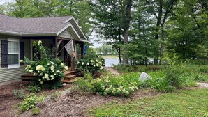 A house with a porch, surrounded by a garden with hydrangeas and a view of a lake. - Fish, Hike & Hunt: Lakeside Newaygo Cottage (Newaygo)